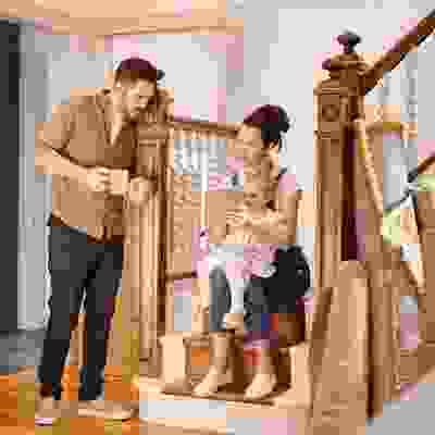 Young family inside a home, with a woman sitting on stairs holding a toddler and a tablet, while a man stands nearby holding two mugs. Rolled-up rugs and moving boxes suggest they are moving in or renovating.