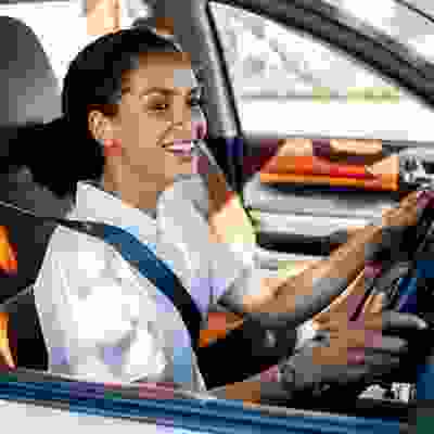 Smiling woman wearing a seatbelt driving a car on a sunny day, looking ahead with her hands on the steering wheel.